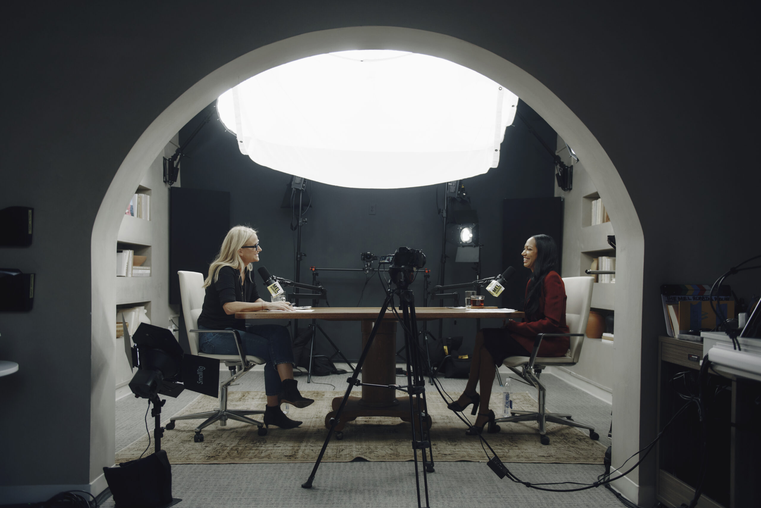 A podcast recording set inside an arched studio space. Two women sit opposite each other at a wooden table, speaking into microphones and smiling. Professional lighting, cameras and audio equipment surround them, with bookshelves built into the walls and a large overhead soft light illuminating the scene.