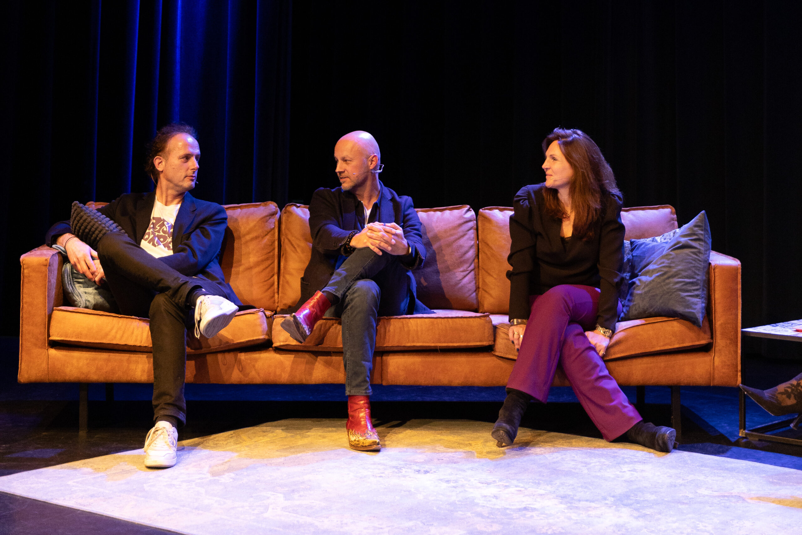 Three people sit on a sofa on a stage, engaged in a panel discussion. They face one another in conversation under warm stage lighting, with a dark backdrop and a relaxed, informal event setting.