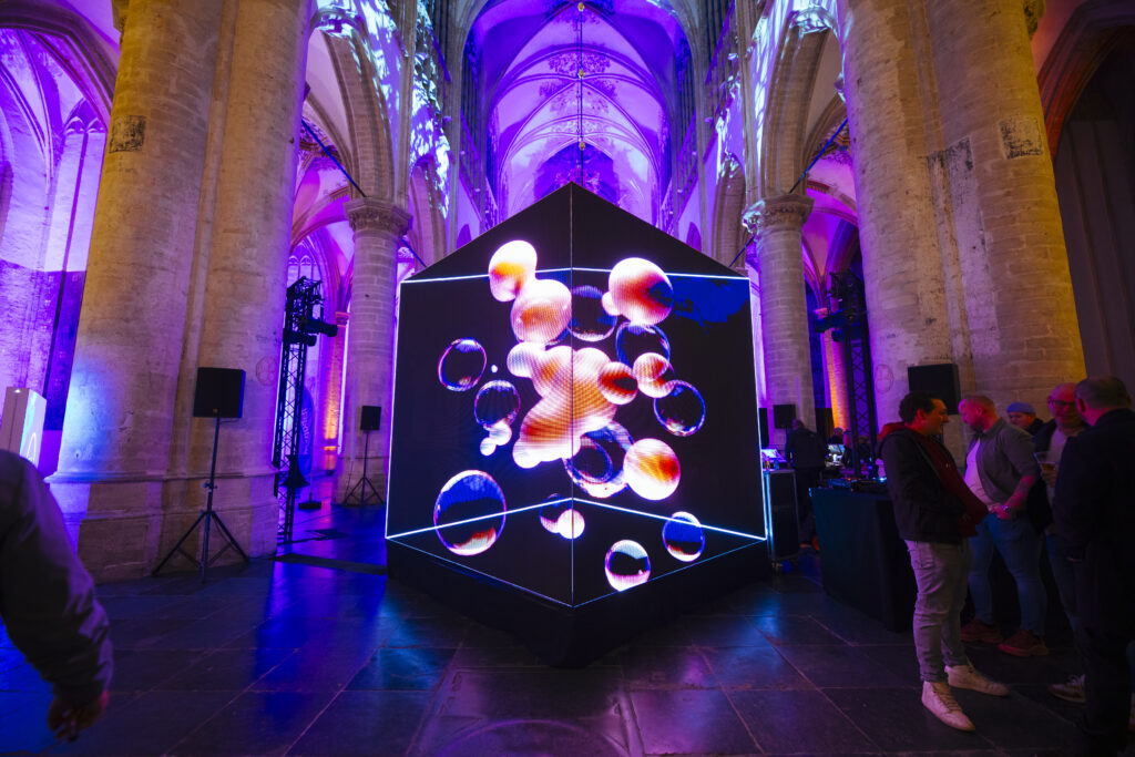 Large digital cube installation glowing with abstract, floating shapes, set inside a historic church with high stone columns and purple lighting, while visitors stand and walk around the artwork.