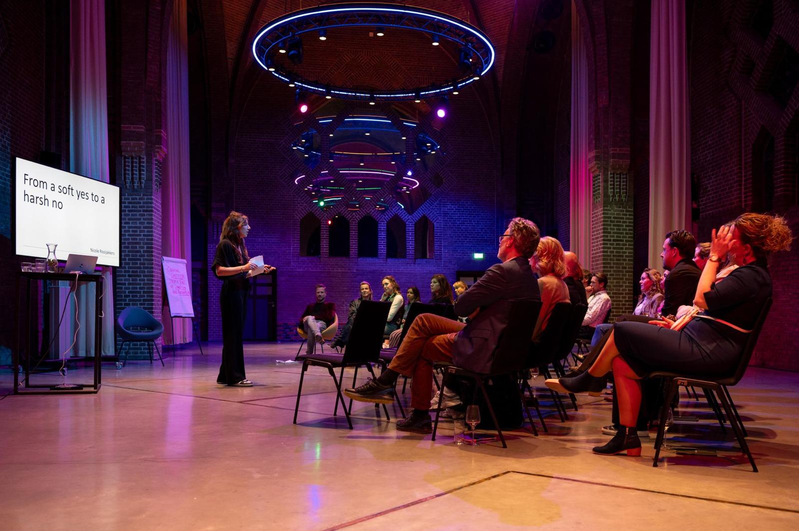 A woman presents to a small seated audience in a spacious church-style venue with high ceilings and brick walls. Coloured stage lighting and circular light installations hang overhead, while a presentation slide reading “From a soft yes to a harsh no” is displayed on a screen to the side.