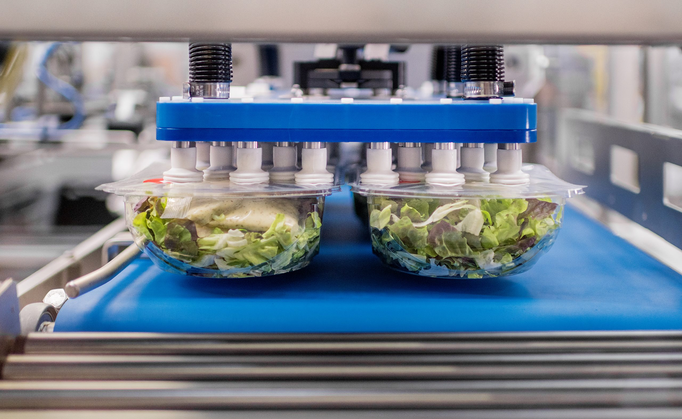 Automated food production line sealing fresh salad bowls in plastic containers at a packaging facility.