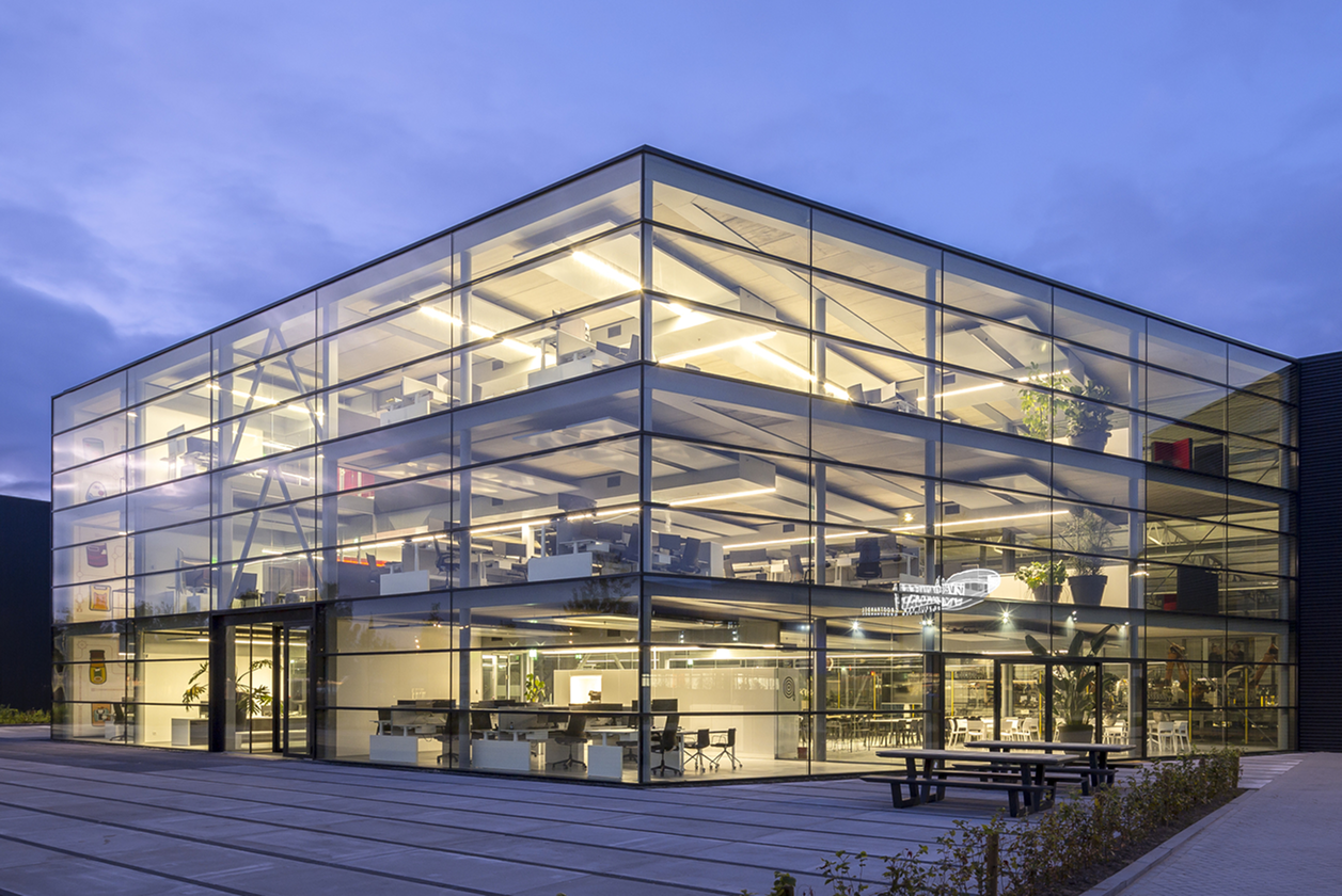 Modern office building with a full glass façade and illuminated workspaces inside, photographed in the evening.